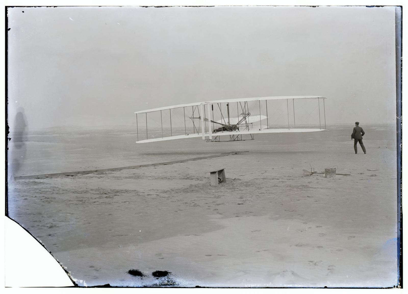 A black-and-white photograph of a fabric biplane just above flat sand, a man lying on its lower wing, while another man in a dark suit stands to the right watching, and a wooden rail runs across the ground.