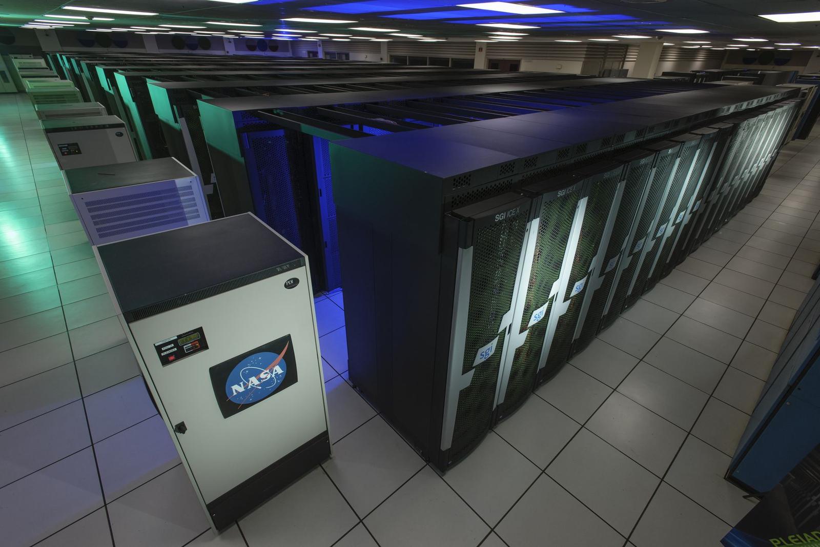 Long rows of tall black computer cabinets glowing blue under ceiling lights in a large tiled room, with a white NASA-branded cabinet in the foreground.